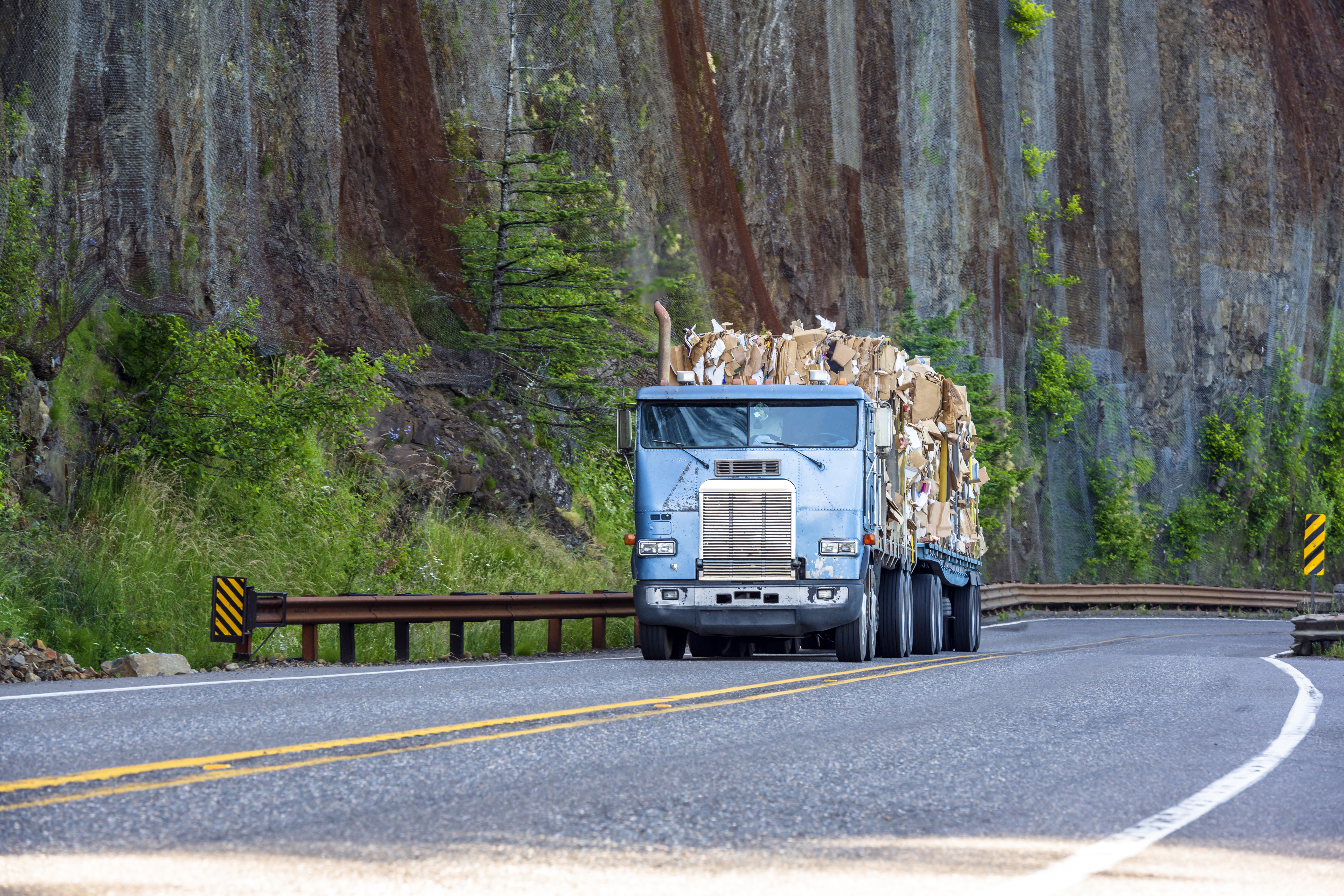Old big rig cab-over semi truck transporting paper recycling on flat bed semi trailer driving on mountain winding road with rock wall.