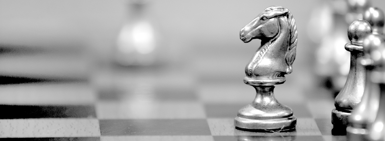 black and white photo of chess pieces on a checkered board with focus on the side profile of a metal horse head representing the knight; side profile of metal pawns align behind the knight