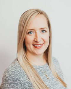 Head and shoulder shot of woman with long blond hair smiling ans wearing a grey knit sweater with a round neckline